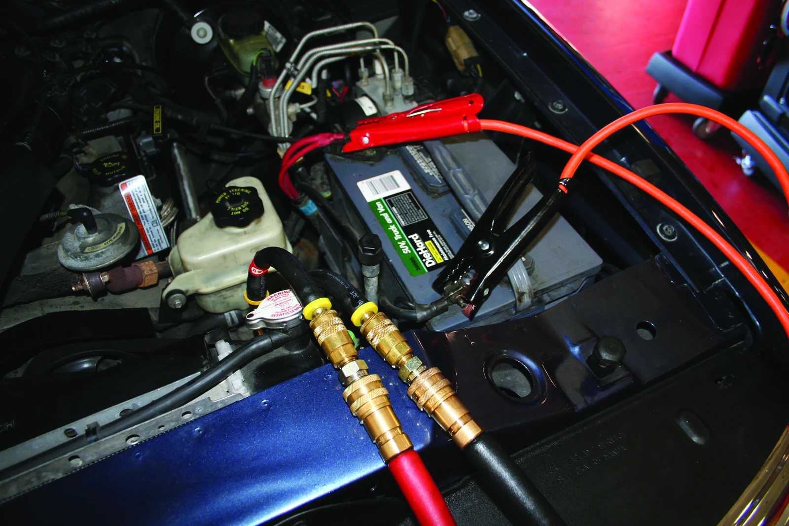 Red and black service hoses joined to transmission cooler-line adapters in a car's engine bay, with power leads connected to the battery